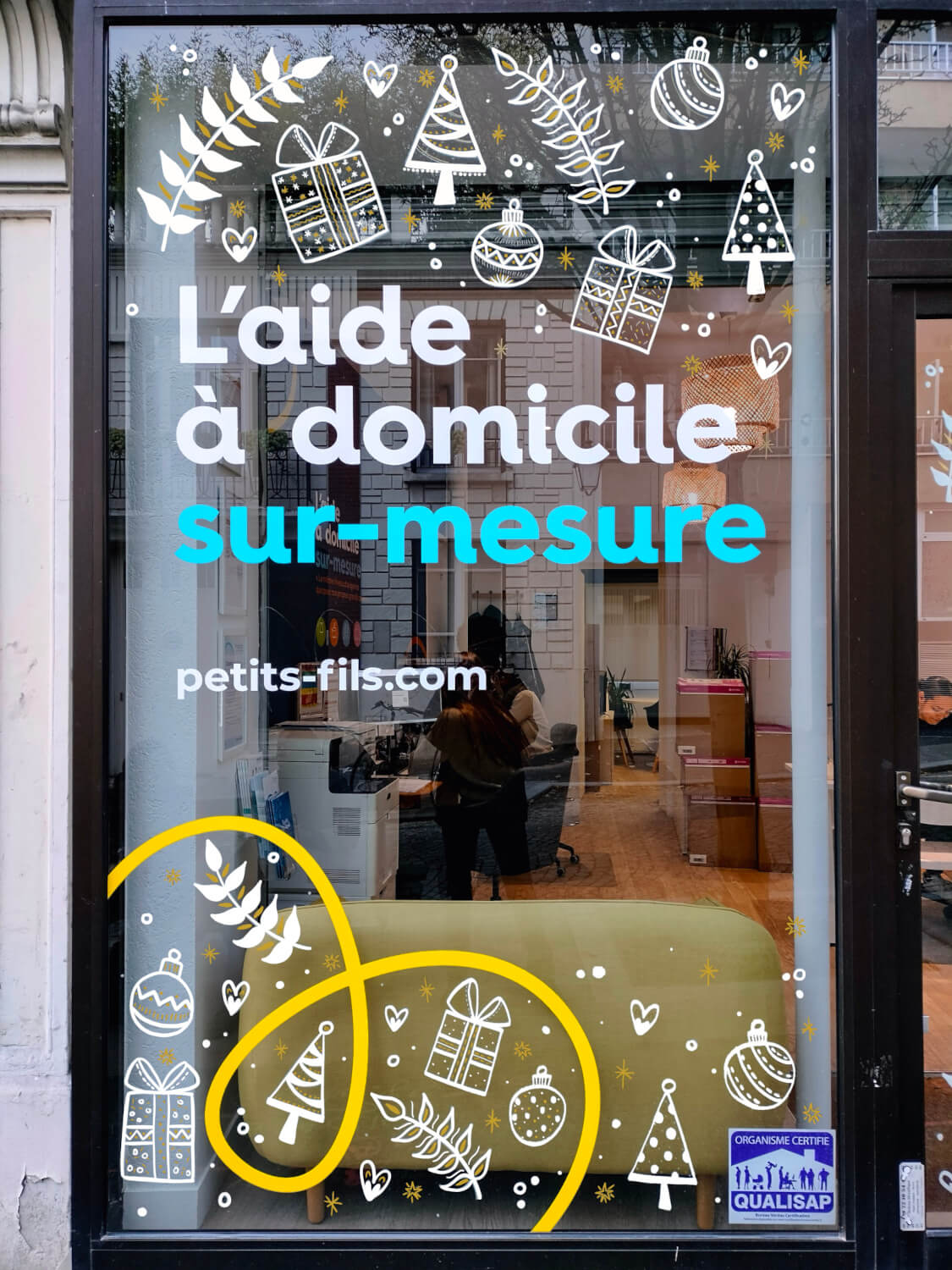 Peinture sur vitrine Paris pour une agence d'aide à domicile sur le thème de Noël en blanc et doré, avec des dessins de cadeaux, sapins, boules de Noël et flocons