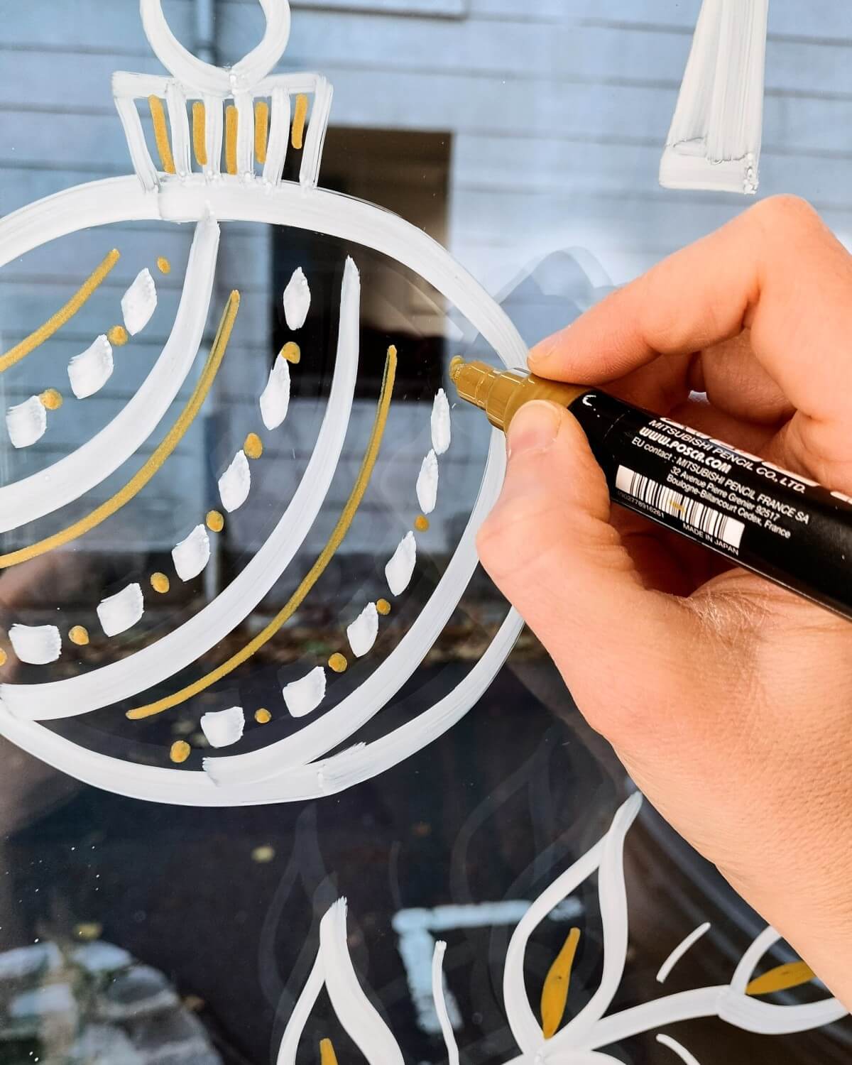 Peinture sur vitrine Rambouillet, détail d'une main qui tient un feutre acrylique Posca et qui dessine une boule de Noël en blanc et en doré